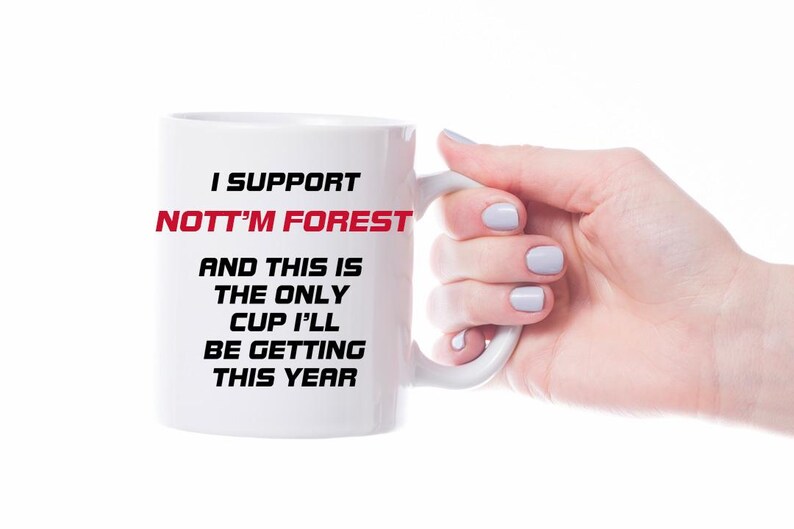 May include: White ceramic mug with the text "I SUPPORT NOTT'M FOREST" in black and red. The text continues with "AND THIS IS THE ONLY CUP I'LL BE GETTING THIS YEAR". The mug is held by a hand with light blue nail polish.