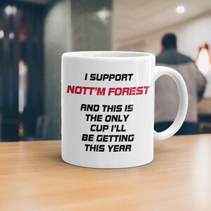 May include: White ceramic mug with the text "I SUPPORT NOTT'M FOREST" in red and black. The mug also reads "AND THIS IS THE ONLY CUP I'LL BE GETTING THIS YEAR". The mug is on a wooden table.