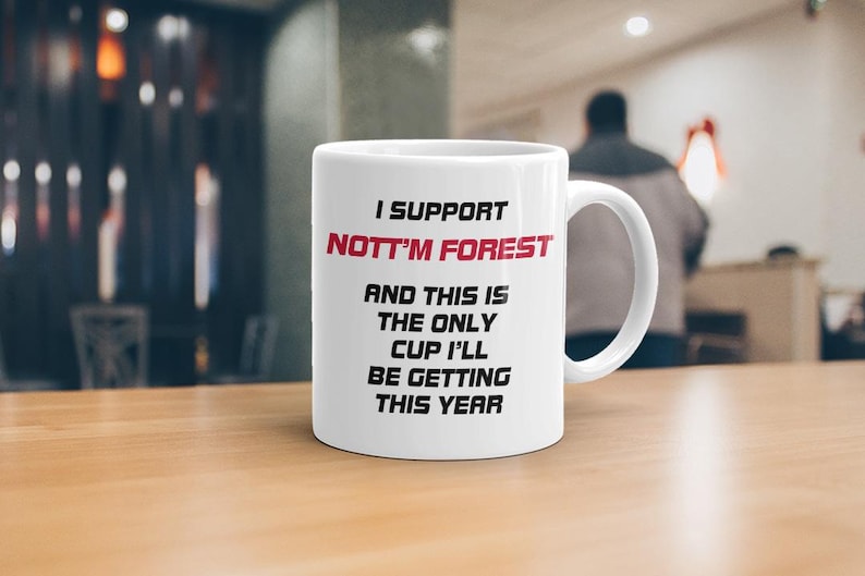 May include: White ceramic mug with the text "I SUPPORT NOTT'M FOREST" in red and black. The mug also reads "AND THIS IS THE ONLY CUP I'LL BE GETTING THIS YEAR". The mug is on a wooden table.