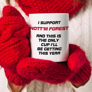 May include: White ceramic mug with the text "I SUPPORT NOTT'M FOREST" in black and red. The mug is held by red knitted mittens. The text continues with "AND THIS IS THE ONLY CUP I'LL BE GETTING THIS YEAR."