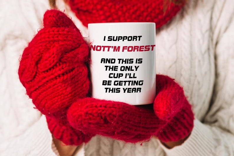 May include: White ceramic mug with the text "I SUPPORT NOTT'M FOREST" in black and red. The mug is held by red knitted mittens. The text continues with "AND THIS IS THE ONLY CUP I'LL BE GETTING THIS YEAR."