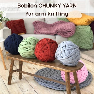 May include: Five balls of chunky yarn in different colors, including navy blue, green, red, gray, and pink, are arranged on a wooden bench. The text "Bobilon CHUNKY YARN for arm knitting" is displayed above the bench.
