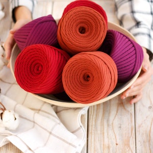May include: A wooden bowl filled with five balls of yarn in shades of red, orange, and purple. The yarn is arranged in a circular pattern, with the ends of the yarn spilling out of the bowl.