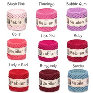 May include: Nine spools of T-shirt yarn in various colors, including blush pink, flamingo, and bubble gum. Each spool has a label that reads "Babilon". The colors range from light pink to deep red and smoky blue.