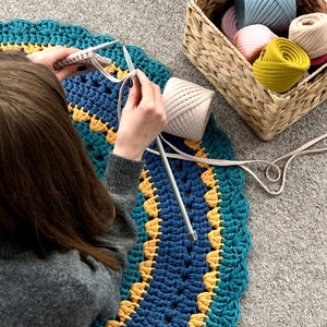May include: A person is knitting a large, round rug with teal, blue, and yellow yarn. A ball of light pink yarn and a basket filled with various yarn colors are nearby. The rug has a scalloped edge and a geometric pattern.