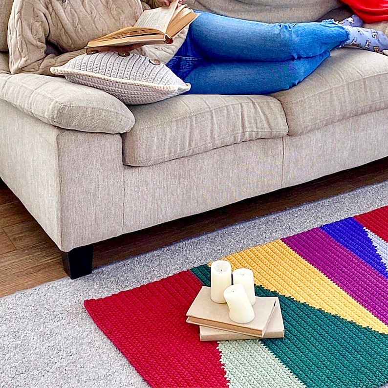 May include: A colorful, handmade crochet rug with a geometric design in red, yellow, green, purple, and blue. Three white candles rest on two books. A person is relaxing on a beige sofa with a crochet pillow.