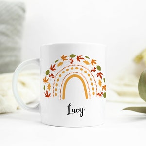 May include: White ceramic mug with a colorful rainbow design featuring autumn leaves and the name "Lucy" printed on the front.