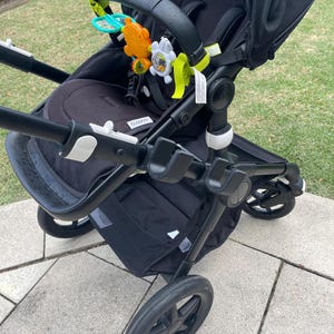 Puede incluir: Un cochecito negro con una capota negra y un asa negra y blanca. El cochecito tiene una cesta negra debajo y un juguete negro y blanco colgando del asa.