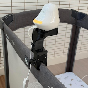 May include: A white, bird-shaped baby monitor with a glowing yellow light base, mounted on a black adjustable clamp attached to the grey fabric frame of a playard. A white power cable is plugged into the side of the monitor and hangs downward.