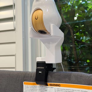May include: A white and light wood-toned baby monitor with a camera lens, mounted on a white plastic stand with a black clamp attached to a grey fabric cot rail featuring a warning label with text about infant safety and mattress usage.