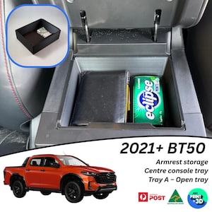 May include: Black armrest storage tray for a 2021+ BT50 truck. The center console tray holds a wallet and a package of Eclipse mints. An additional small black tray is shown in the upper left corner. The text reads "Armrest storage, Centre console tray, Tray A - Open tray."