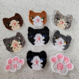 May include: Nine crocheted cat faces and two paw prints in various colours, including brown, grey, black, and white. The cat faces have white yarn for the eyes and nose.