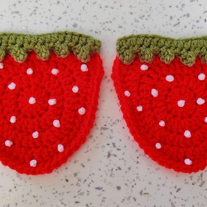 May include: Two strawberry-shaped crochet coasters made with bright red yarn. Each coaster features a textured green crochet border at the top to represent leaves and small white yarn knots scattered across the surface to mimic strawberry seeds.