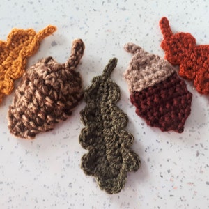 May include: Five crocheted autumn leaves and acorns in various colours, including brown, green, orange, and red.