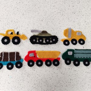 May include: Six crocheted toy vehicles are displayed, including a yellow excavator, a black and green tank, a yellow dump truck, a blue truck with brown logs, a red dump truck, and a green truck. Each vehicle has black wheels.