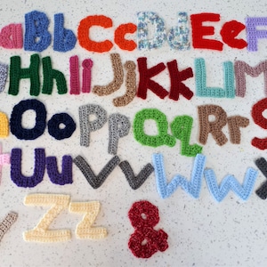 May include: A set of crocheted letters in various colours, including pink, green, blue, red, yellow, and purple. The letters are arranged in alphabetical order, with the letter 'A' at the top left and the letter 'Z' at the bottom right. There is also a crocheted ampersand.