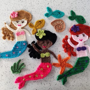 May include: A collection of crocheted mermaid and sea creature appliques. The mermaids have brown, black, and red hair, with pink and blue tails. Other elements include fish, a starfish, and seaweed, all in vibrant colours.