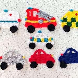 May include: A set of seven crocheted toy cars in various colours, including red, blue, yellow, green, and white. The cars have black wheels and are decorated with details like headlights, windscreens, and sirens.