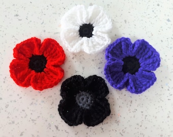 May include: Four handmade crochet flower appliques arranged in a cluster. The set includes one red flower with a black centre, one white flower with a black centre, one purple flower with a black centre, and one black flower with a dark grey centre.