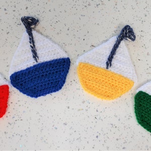 May include: Four crocheted sailboats in different colours. The sailboats are white with a coloured bottom. The colours are red, blue, yellow, and green.