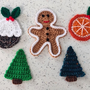 May include: A selection of crocheted Christmas decorations. The image shows a Christmas pudding, a gingerbread man, an orange slice, and two Christmas trees. The ornaments are crafted from yarn in various colours and are displayed on a speckled surface.