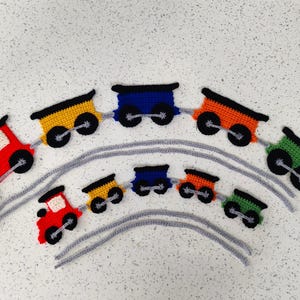 May include: A crocheted train set in red, yellow, blue, orange, and green. Each train carriage has black wheels and a black roof. The carriages are connected by grey yarn, creating a playful and colourful design.