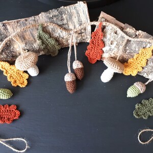 May include: A handcrafted autumn garland with crocheted leaves, mushrooms, and acorns in orange, yellow, green, and brown. The garland is strung on twine and displayed against a birch bark backdrop, perfect for seasonal decor.