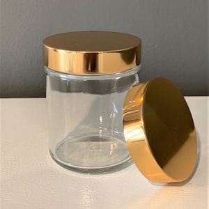 Op de afbeelding: Een glazen pot met een gouden deksel. Het deksel is eraf en ligt naast de pot.