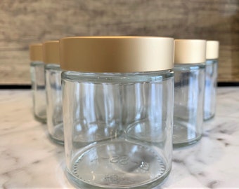 May include: Five clear glass jars with gold lids. The jars are arranged in a row on a white surface.