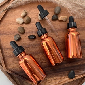 May include: Three copper colored glass bottles with black dropper tops on a wooden tray. The bottles are filled with a clear liquid.