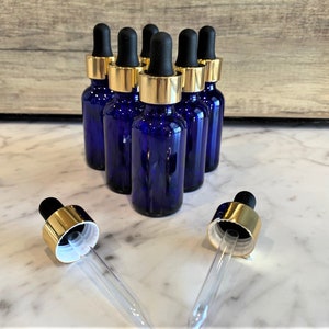 May include: Six cobalt blue glass bottles with gold lids and black droppers. The bottles are arranged in a row on a white marble surface.