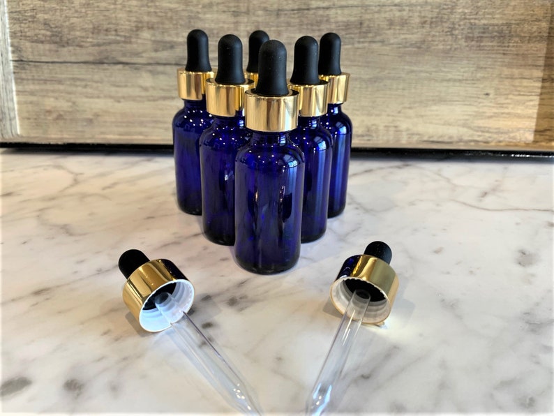 May include: Six cobalt blue glass bottles with gold lids and black droppers. The bottles are arranged in a row on a white marble surface.