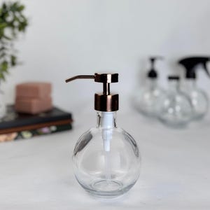 May include: A clear, round glass soap dispenser with a copper-colored pump. The dispenser has a white plastic tube and is sitting on a white surface. Other similar dispensers are blurred in the background.