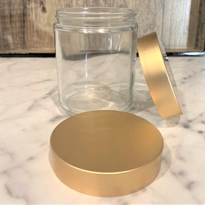 May include: A clear glass jar with a gold lid. The lid is off the jar and sitting on a white marble surface.