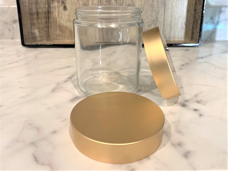 May include: A clear glass jar with a gold lid. The lid is off the jar and sitting on a white marble surface.