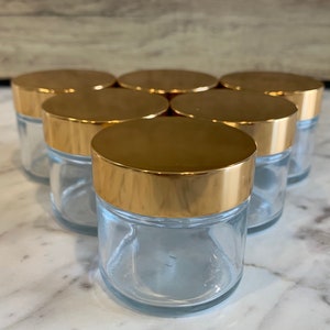 May include: Set of six clear glass jars with gold lids. The jars are arranged in a circle with the lids facing up.