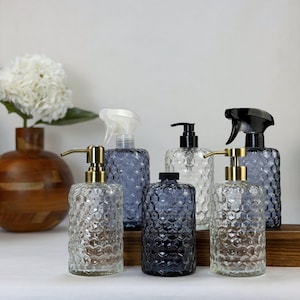 May include: A collection of five glass bottles with a hexagonal honeycomb texture. Two bottles feature gold-toned pump dispensers, one has a black pump, one has a white spray nozzle, and one has a black spray nozzle. The bottles vary in clear and dark blue glass.