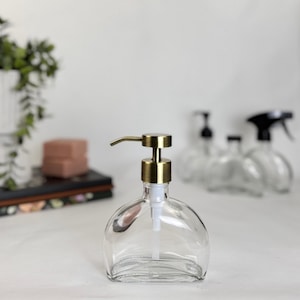 May include: Clear glass soap dispenser with a brass-colored pump. The dispenser has a rounded shape and a white pump tube. Other clear glass bottles are visible in the background.