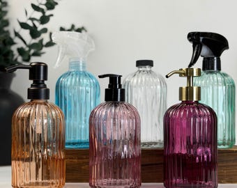 May include: A collection of decorative glass bottles in various colors, including amber, pink, clear, and teal. Some bottles have pump dispensers, while others have spray nozzles. The bottles are arranged on a wooden surface.