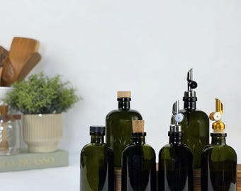 May include: A collection of six dark green glass bottles of varying heights. Some bottles feature black screw caps, others have natural cork stoppers, and two are topped with silver or gold-colored metal pour spouts for dispensing liquids.