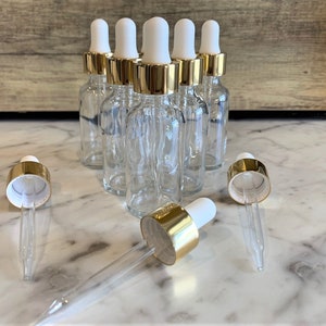 May include: Clear glass dropper bottles with gold-colored collars and white tops. The bottles are arranged in a group, with several droppers lying in front. The bottles are empty and appear to be for essential oils or cosmetic products.