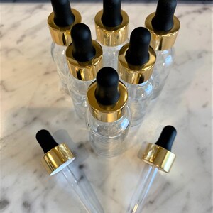 Op de afbeelding: Acht heldere glazen flessen met gouden deksels en zwarte druppelaars. De flessen zijn in een cirkel gerangschikt met twee flessen die op hun zij liggen.