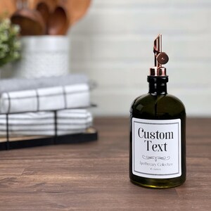 Pu&ograve; includere: Una bottiglia di vetro verde scuro con un tappo a pompa in rame e un'etichetta bianca che dice "Custom Text" e "Apothecary Collection 16 ounces".