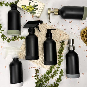 Op de afbeelding: Een verzameling zwarte flessen met verschillende dispensers, waaronder sproeiers en pompen. Sommige flessen hebben een lichtgroene of grijze basis. Er is ook een blok olijfoliezeep aanwezig, samen met decoratief groen.