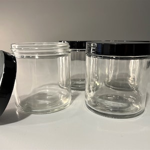 Puede incluir: Cuatro tarros de vidrio transparente con tapas negras. Los tarros están vacíos y tienen forma redonda.
