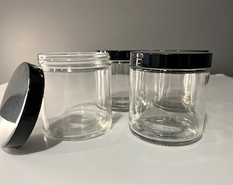 May include: Four clear glass jars with black lids. The jars are empty and have a round shape.
