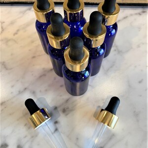 May include: Seven cobalt blue glass bottles with gold lids and clear glass droppers. The bottles are arranged in a pyramid shape on a white marble surface.