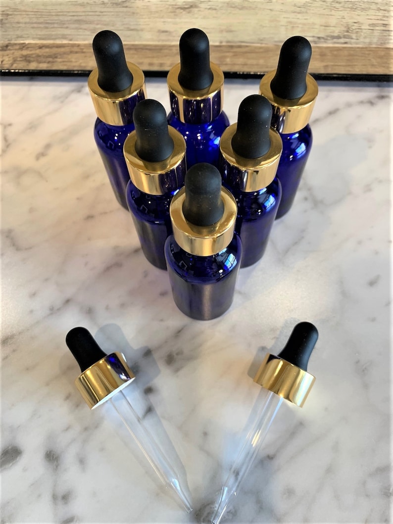 May include: Seven cobalt blue glass bottles with gold lids and clear glass droppers. The bottles are arranged in a pyramid shape on a white marble surface.