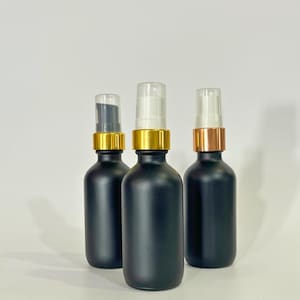 Könnte beinhalten: Drei schwarze Glasflaschen mit goldenen und kupferfarbenen Verschlüssen. Die Flaschen haben transparente Pumpen.