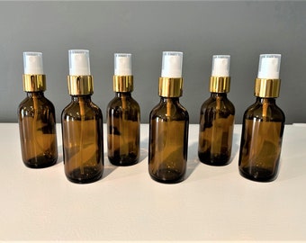 Könnte beinhalten: Sechs bernsteinfarbene Glasflaschen mit goldenen Deckeln und weißen Sprühköpfen. Die Flaschen sind in einer Reihe auf einer weißen Oberfläche angeordnet.